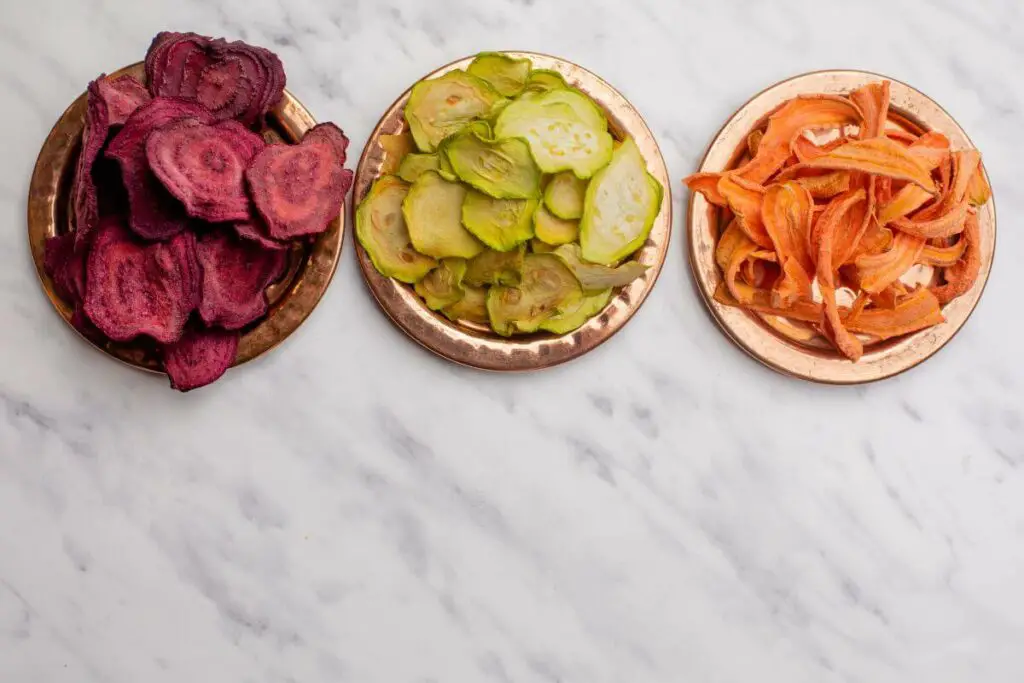 Three bowls of snacks, including carrots, beets, and radishes, are beautifully arranged on a marble table.