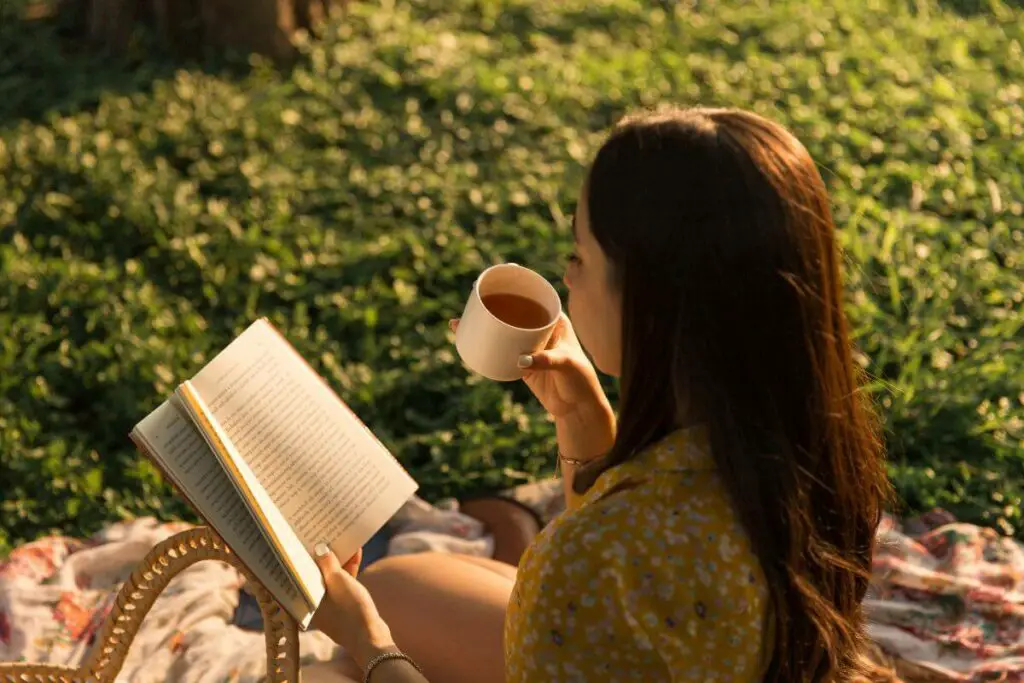A woman is sitting on the grass reading a book and drinking a cup of coffee, unaffected by any concerns related to vegetables and IBS.