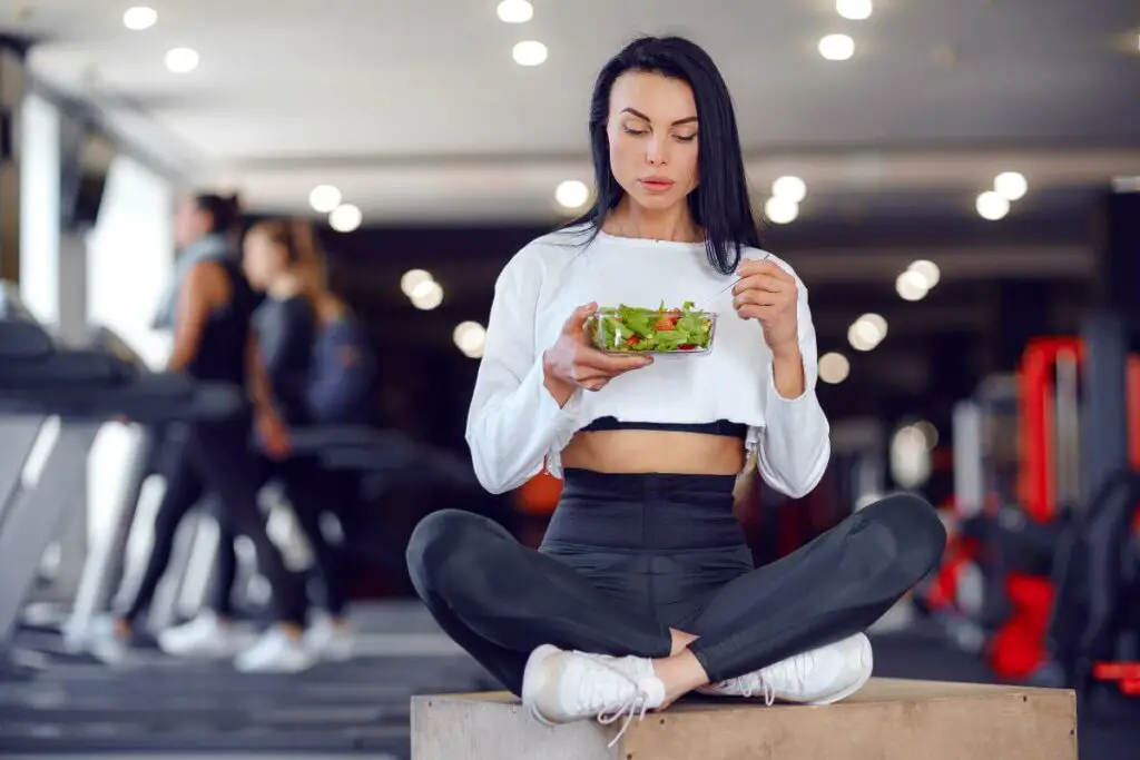 A woman enjoying a raw vegetable salad in a gym.