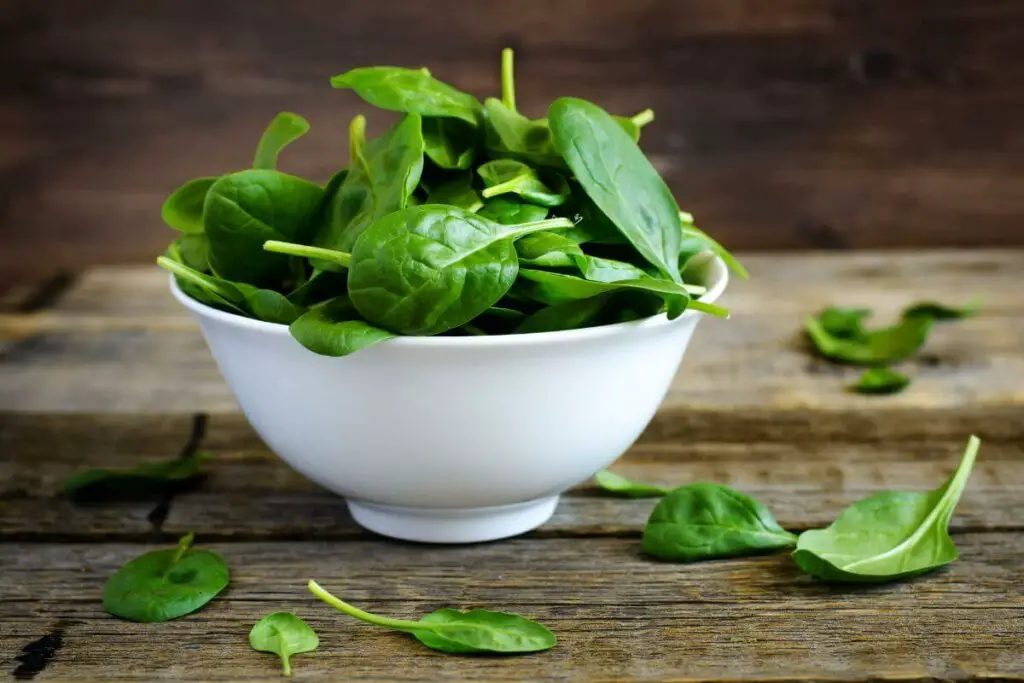 Spinach leaves in a white bowl on a wooden table, suitable for individuals with irritable bowel syndrome (IBS) and looking to incorporate vegetables into their diet.