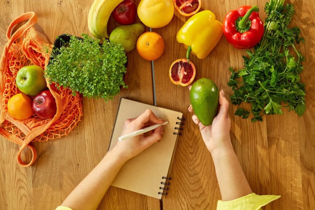 A woman writing on a notebook next to a farmer's market stall with fruits and vegetables.