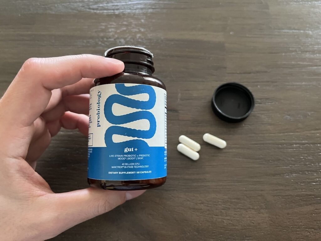A hand holds a brown bottle labeled “probiotic gut+” with a blue and white design. The black lid is off and on the table, next to three white capsules, evoking images from a typical yourbiology gut+ review.