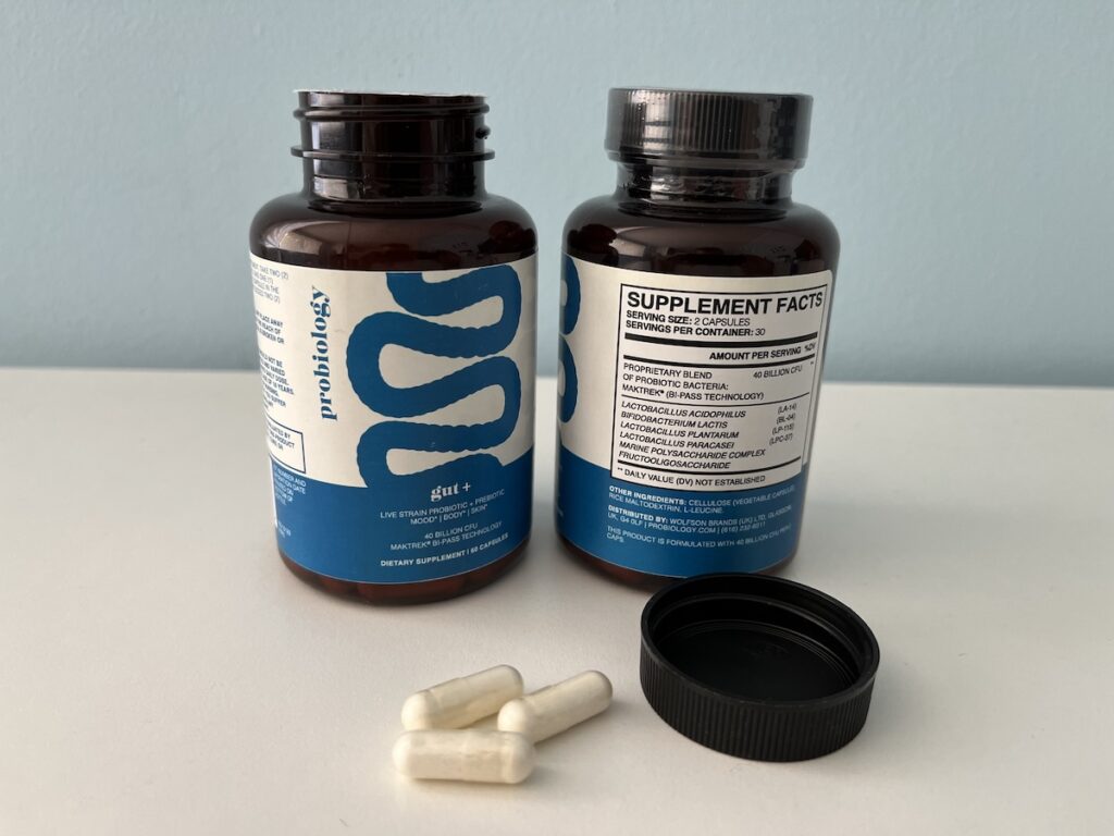 Two brown bottles labeled “probiology gut+” with blue and white labels are displayed side by side. One bottle reveals the supplement facts, while three white capsules and a black bottle cap rest on the white surface in front of them—ideal for readers seeking a detailed yourbiology gut+ review.